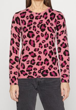 Femme portant un pull rose à manches longues avec un imprimé léopard noir et rose foncé et un pantalon gris foncé, debout devant un fond uni.