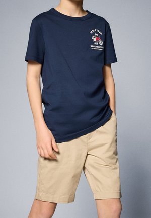 Dziecko ubrane w granatowy T-shirt z małym logo motocykla oraz beżowe szorty, stojące na jednolitym szarym tle.