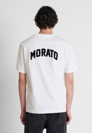 Persona con capelli ricci e scuri che indossa una maglietta bianca con testo nero "MORATO" sulla schiena e jeans neri, rivolta di spalle alla fotocamera.