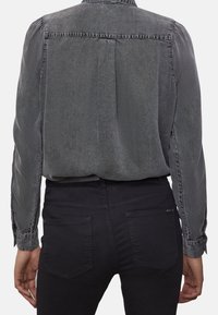 Person som bär en tvättad svart denimskjorta med lång ärm, instoppad i högmidjade svarta jeans, sedd bakifrån.