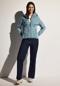 Giacca puffer azzurro chiaro con cappuccio rimovibile, chiusura con zip e tasche frontali. Indossata con jeans scuri e sneakers chiare.