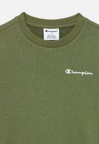 Πράσινο ελιάς μπλουζάκι από απαλό ύφασμα, με στρογγυλή λαιμόκοψη, κοντά μανίκια και λευκό λογότυπο «Champion» στο στήθος.