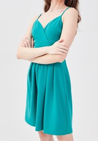 Robe sans manches de couleur sarcelle avec un corsage ajusté et une jupe évasée. Texture lisse avec un décolleté en V et un détail de taille froncée.