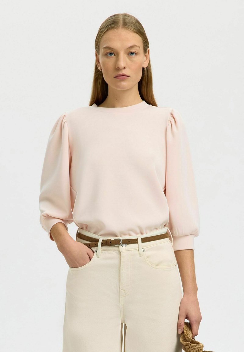 Femme aux longs cheveux blonds portant un haut rose pâle à manches bouffantes, un pantalon crème taille haute avec ceinture marron, tenant un sac tressé marron.