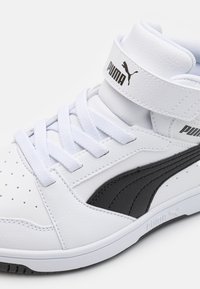Baskets montantes en cuir blanc avec des accents noirs, des sections perforées, des lacets plats et une sangle Velcro portant le logo Puma.