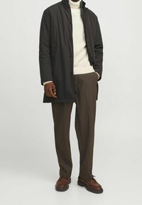 Homme portant un manteau noir boutonné par-dessus un col roulé blanc en maille torsadée, un pantalon marron et des chaussures en cuir marron, debout avec une main dans la poche.