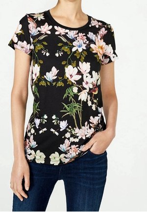 Kobieta ubrana w czarną koszulkę z krótkim rękawem o wielokolorowym, kwiatowym wzorze i wzorem ważek, w połączeniu z ciemnoniebieskimi dżinsami.