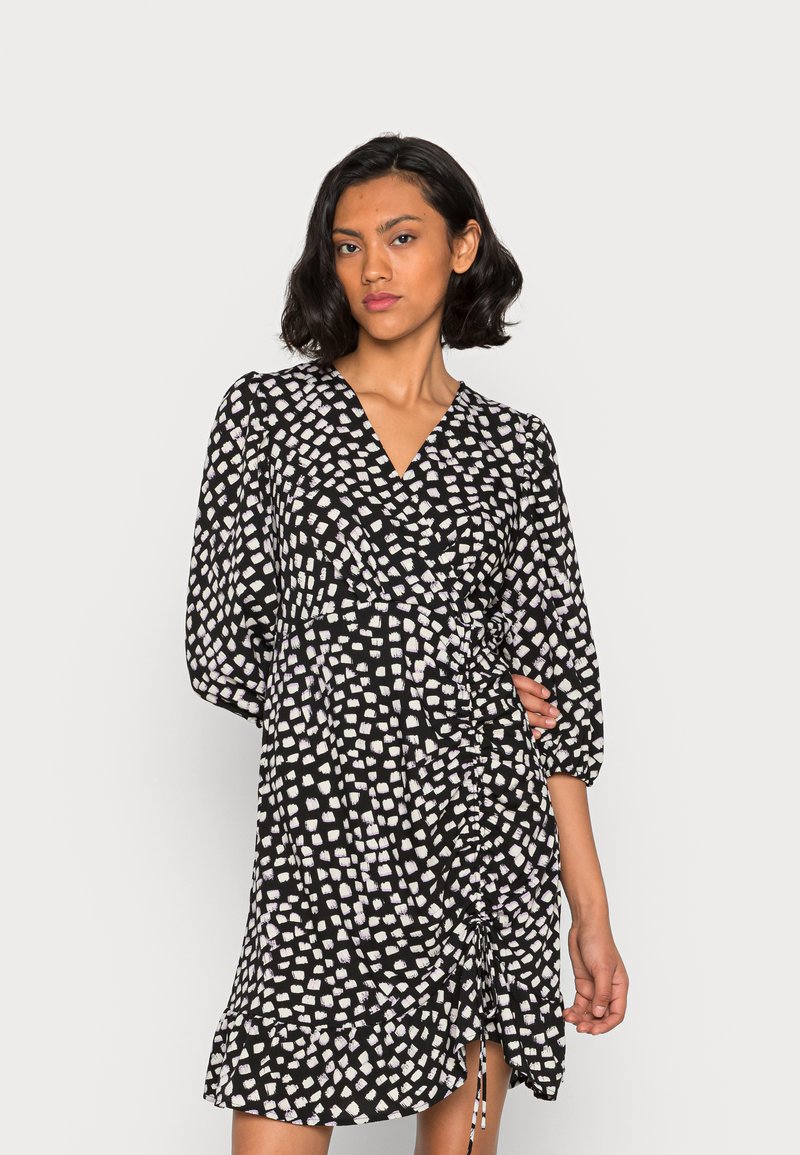 Vestido preto em envoltório com um padrão quadrado branco, decote em V, mangas três quartos, franzido lateral e detalhe de cordão. Textura de tecido suave.