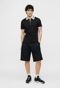 Νεαρός άνδρας φορώντας μαύρο κοντομάνικο πόλο με λευκό γιακά και μανσέτες με λογότυπο, μαύρα σορτς, λευκές κάλτσες και μαύρα αθλητικά παπούτσια, στέκεται με τα χέρια στις τσέπες.