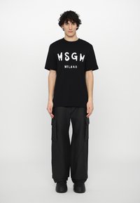 Musta puuvillapaita, jossa on valkoinen "MSGM MILANO" -painatus, yhdistettynä löysiin mustiin cargo-housuihin, joissa on sivutaskut ja sileä pinta.