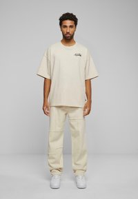 Beige oversized t-skjorte med rund hals og frontlogo, kombinert med matchende beige vide bukser med lappeteknikk. Hvite sneakers.