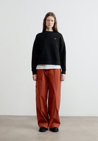 Όρθιο άτομο με μακριά μαλλιά που φοράει μαύρο φούτερ, λευκό εσώρουχο, φαρδιά παντελόνια cargo σκουρόχρωμου σκουριάς και μαύρα παπούτσια, σε απλό φόντο.