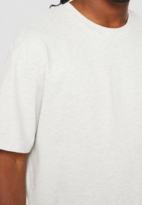 Světle šedé tričko s krátkým rukávem vyrobené z měkké, texturované tkaniny. Má kulatý výstřih a volný střih. Žádné výrazné vzory ani akcenty.