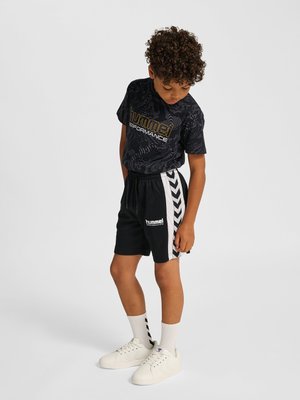 Jongen met krullend haar, die een zwart T-shirt met patroon draagt en zwarte sportshorts met witte chevronstrepen, kijkt naar zijn shorts.