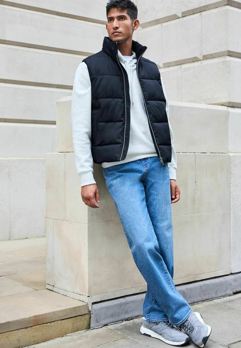 Zwarte gewatteerde vest over een lichtgrijze hoodie, gecombineerd met blauwe jeans en grijze sneakers. Achtergrond bestaat uit neutrale stenen architectuur.