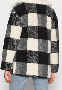 Schwarz-weiße und graue kariert Fleecejacke mit einem weichen Kragen, getragen von einer Person in hellblauen Jeans, von hinten gezeigt.