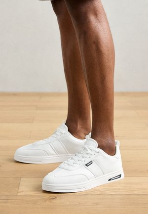 Persona che indossa sneakers basse bianche con il marchio nero "MORATO" e "ANTONMORATO", in piedi su un pavimento in legno chiaro.