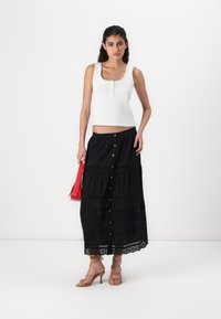 White ribbed top with square neckline and buttons, paired with a black ankle-length skirt featuring layered panels and lace trim. Red handbag and strappy beige heels.