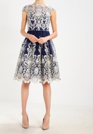 Vestido navy con un sobrecosto de encaje en crema. Mangas cortas, corpiño ajustado y falda acampanada con detalles de dobladillo en forma de ondulaciones.