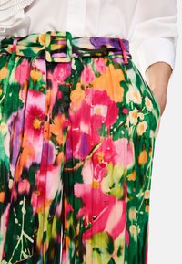Faltende Blumenhose mit lebendigen Pink-, Grün- und Gelbtönen; verfügt über einen breiten grünen Quadratschnalle und seitliche Taschen. Leichter Stoff.