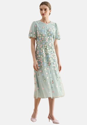 Frau trägt ein grünes Midikleid mit Puffärmeln und pastellfarbener Blumenstickerei, kombiniert mit hellrosa flachen Schuhen, stehend vor weißem Hintergrund.