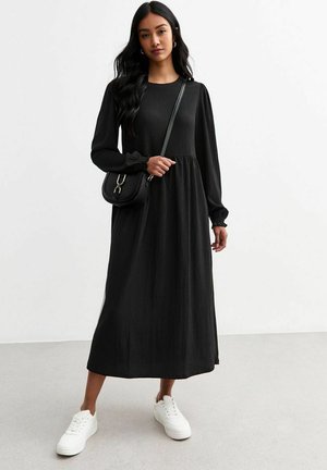 Zwart lange mouwen jurk met een gestructureerde stof, gathered taille en een losse pasvorm. Gestyled met witte sneakers en een kleine zwarte schoudertas.