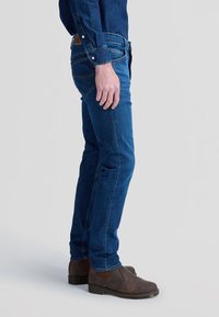Homme portant un jean bleu foncé, des bottes en cuir marron et une chemise en denim bleu foncé, debout devant un fond uni.