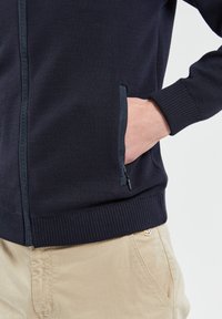 Veste en tricot bleu marine à fermeture éclair, avec des poignets et un ourlet côtelés. Dispose d'une poche latérale zippée. Portée par-dessus un pantalon beige avec un détail de logo.