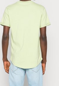 Personne portant un T-shirt vert clair à manches courtes et un jean bleu clair, vue de dos sur un fond uni.