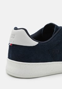 Talons de baskets en daim bleu marine avec col matelassé blanc, semelle blanche tachetée et petit détail en tissu rouge, blanc et bleu au dos.