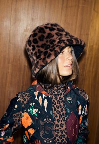 Pălărie de tip bucket cu imprimeu leopard, realizată din blană artificială moale, având o borură lată. Modelul poartă o jachetă cu modele multiple și culori vibrante.