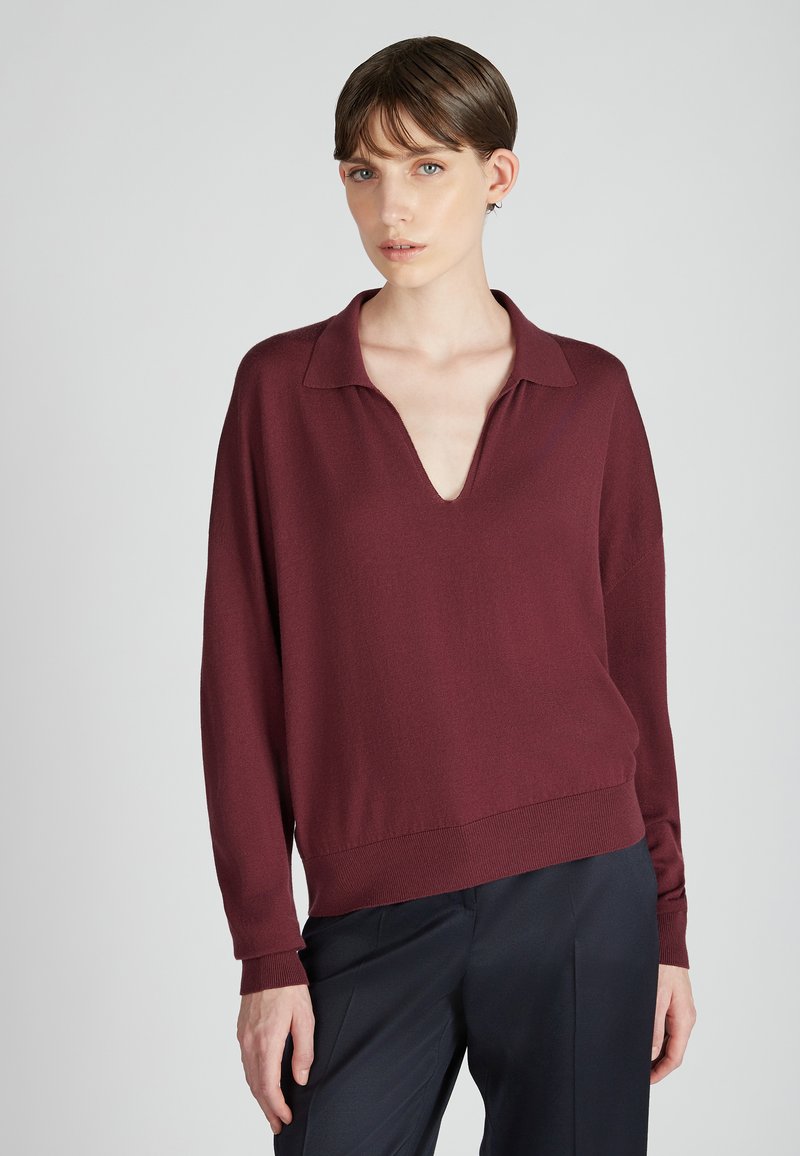 Burgundowy pulower z długim rękawem, głębokim dekoltem w serek i ściągaczem na dole; miękka tekstura; noszony z ciemnymi, klasycznymi spodniami.