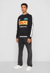 Svart sweatshirt med färgglada logotyper, vita detaljer på ärmarna, avslappnad passform, matchad med mörka denimjeans och vita sneakers.