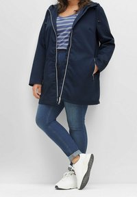 Donkerblauw gewatteerd jas met capuchon en witte ritssluiting, met een rechte snit en twee zijzakken, gedragen over een gestreept shirt en blauwe spijkerbroek. Witte sportieve sneakers.