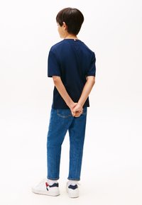 Boy with short brown hair wearing a navy t-shirt, blue jeans, white sneakers, standing with hands clasped behind back on white background.