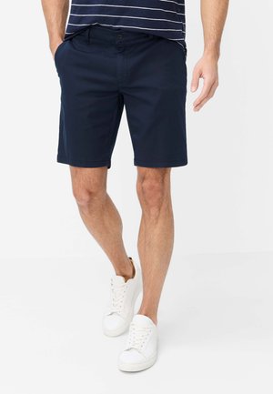 Mand iført mørkeblå shorts, hvide sneakers og en navyblå T-shirt med striber, stående med den ene hånd i lommen mod hvid baggrund.