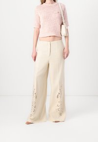 Hellrosa, kurz geschnittenes, strukturiertes Strickoberteil mit kurzen Ärmeln und einer kleinen Tasche. Beigefarbene weit geschnittene Hose mit dekorativen Ausschnitten.