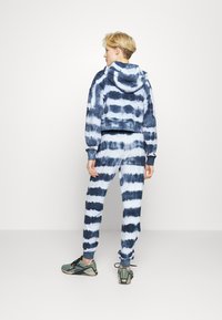 Cropped hoodie azul tie-dye com calças de jogger combinando, apresentando riscas horizontais, punhos elásticos e um bolso canguru. Usado com ténis.