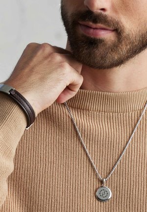 Silver pendant necklace with a circular charm featuring a compass design. Worn over a textured tan sweater. Brown leather bracelet on wrist.