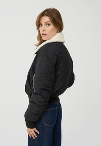 Czarna bomberka z białym futrzanym kołnierzem, zapinana na zamek, z elastycznymi mankietami i kieszenią z boku. Noszona z niebieskimi dżinsami.