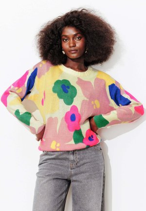 Femme aux cheveux bouclés volumineux portant un pull coloré à fleurs et un jean gris, posant les mains sur les hanches devant un fond blanc.