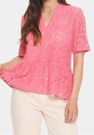 Femme portant un chemisier court à manches courtes en dentelle florale rose associé à un pantalon beige clair, tenant un côté de l'ourlet du chemisier.