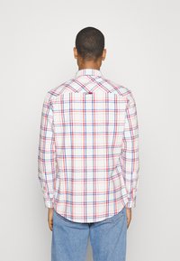 Man med långärmad vit skjorta med blå och röd rutmönster, stående vänd bort, ihop med ljusblå jeans mot en enfärgad bakgrund.