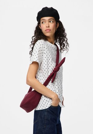T-shirt bianco con pois neri e maniche corte, abbinato a jeans blu scuro e una borsa a tracolla bordeaux. La borsa ha una tracolla annodata e una superficie liscia.