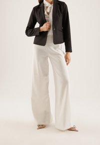 Black tailored blazer with a single button, white blouse with a neck tie, and wide-leg white trousers. The shoes are nude with bow accents.