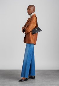 Kiilane inimene seistes risti käed rinnal, seljas pruun nahkblazer, triibuline kõrge kaelusega särk, laiad sinised teksad, mustad lamedad kingad ning väike must õlakott.