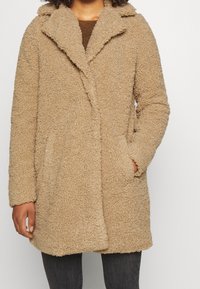 Beige kabát s chlupatou texturou, límcem, dvěma bočními kapsami a střední délkou. Měkký, teplý materiál vhodný pro chladnější počasí.