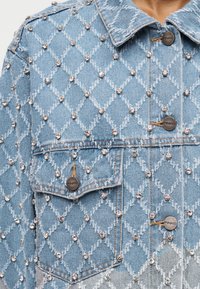 Φωτεινό μπλε τζιν μπουφάν με ραφές σε σχήμα διαμαντιού και μικρές στρογγυλές ασημένιες καρφίτσες, με μεταλλικά κουμπιά και τσέπη στο στήθος.