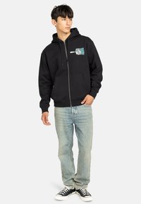 Felpa nera con zip e tasca frontale, decorata con un design grafico sul lato sinistro del petto. Abbinata a jeans chiari e sneakers nere.