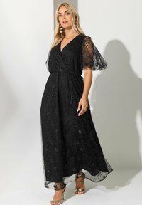 Musta pitsinen maksimekko lyhyillä leijonasateilla, monimutkaisilla brodeeratuilla kukkakuvioilla ja kapealla vyötäröllä, yhdistettynä hihnakenkiin.